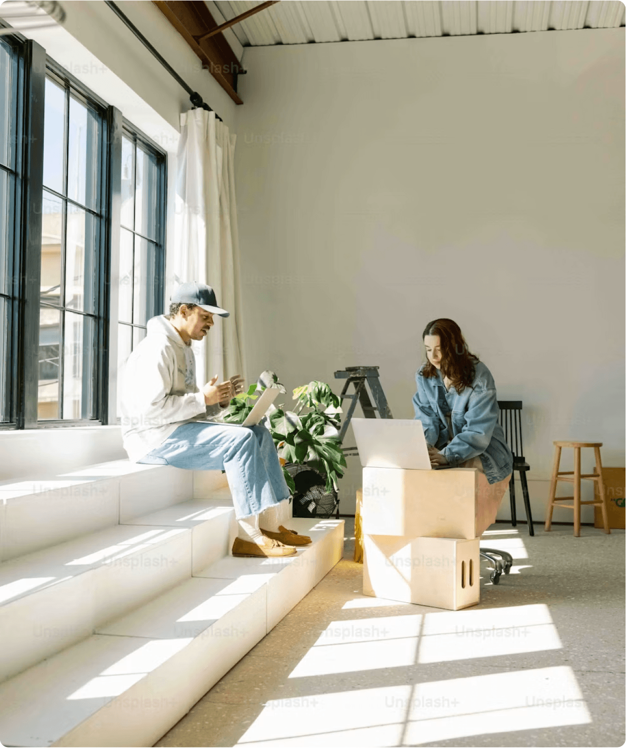 Two people working on laptops beside a sunlit window.